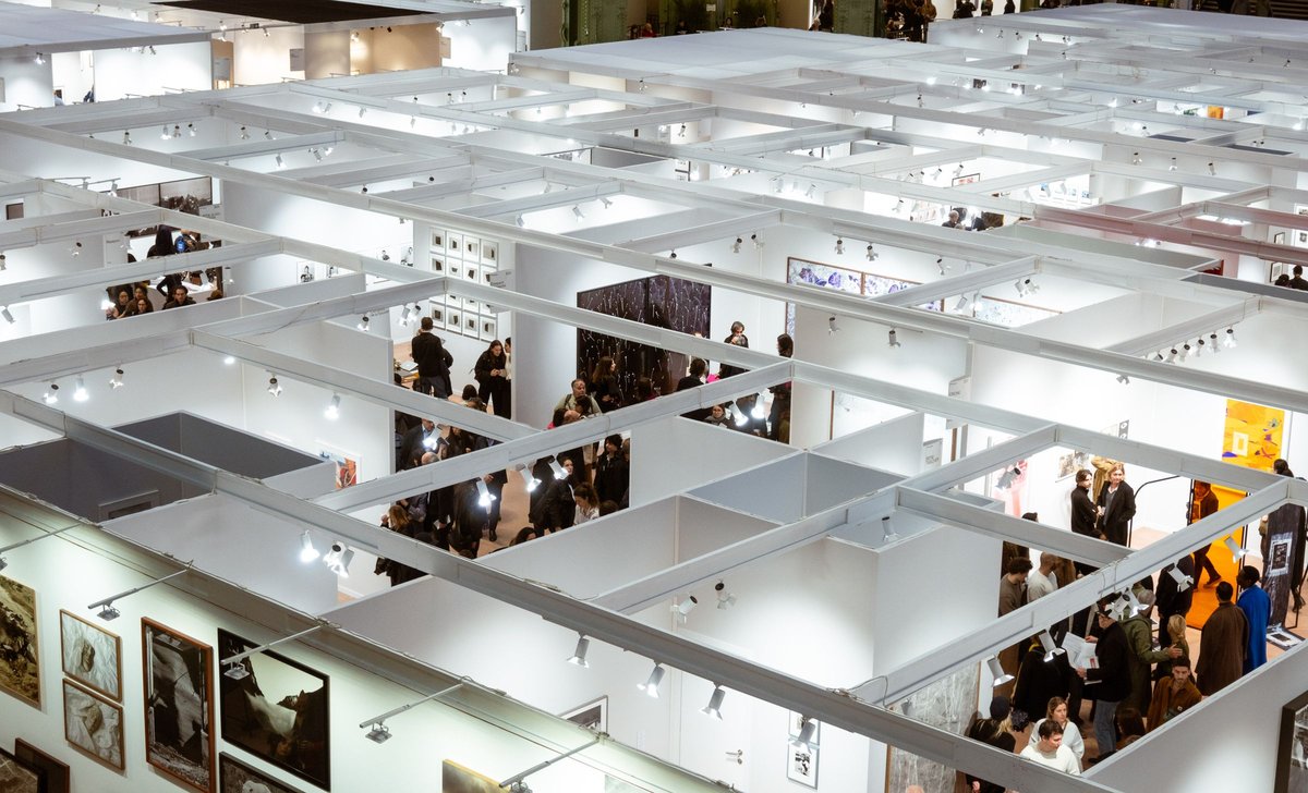 Aerial view of a large exhibition hall divided into a grid of open-topped booths, with visitors walking between them.