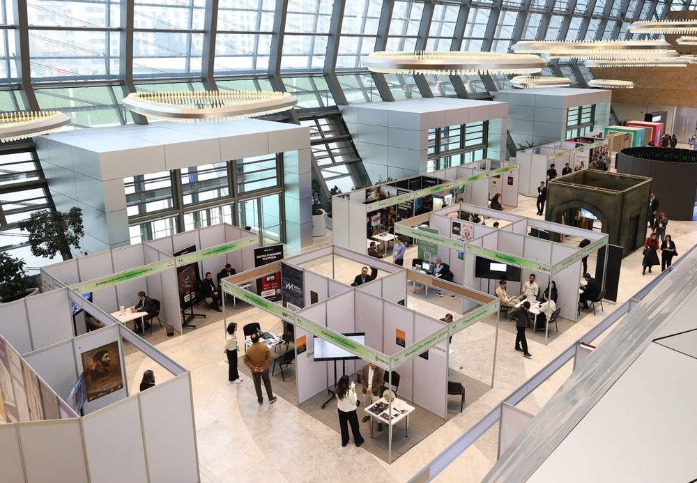 Aerial view of a modern glass-roofed exhibition hall with small booths and hanging light fixtures.